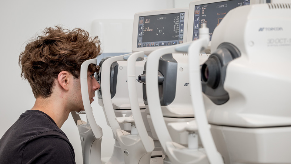 A young man is positioned at a device for a sight test. Ahead of him is a row of several more testing devices