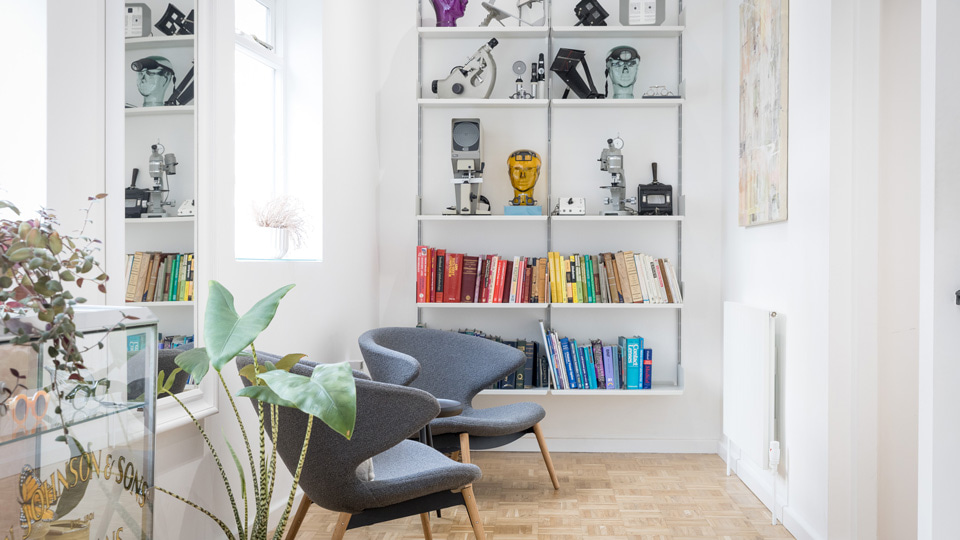 A waiting space with two comfortable, modern grey chairs, potted plants, and a display case with two rows of books arranged in a rainbow colour, as well as models of heads wearing optometric equipment, as well as optical technology