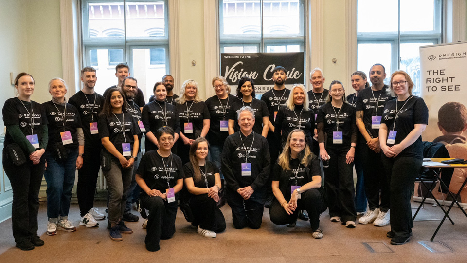Group photo of around 20 volunteers wearing black OneSight EssilorLuxottica Foundation T-shirts, posing inside a bright room with tall windows. Some stand while others kneel at the front, all smiling at the camera. Behind them, a large sign reads “Welcome to the Vision Clinic,” with additional branding for “The Right to See” visible on a banner to the right