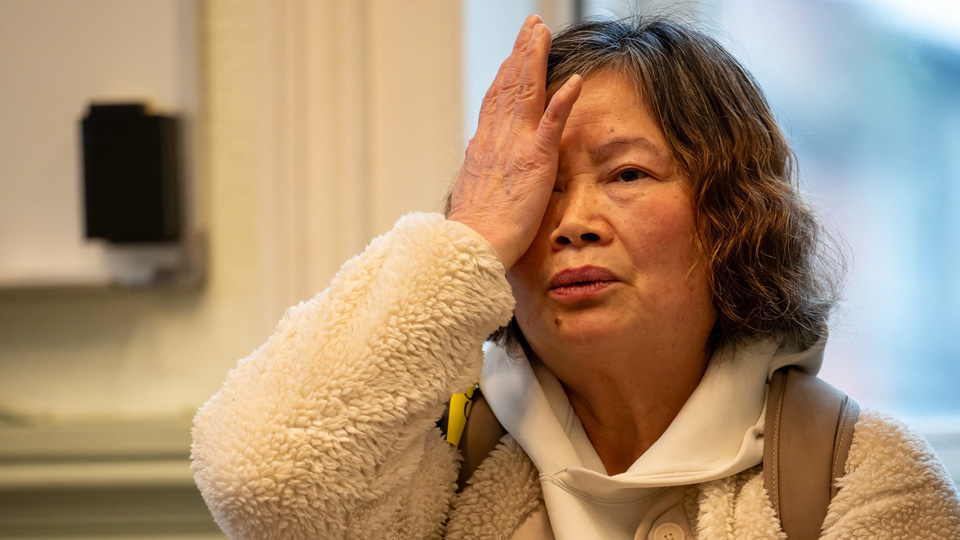 An older woman sits indoors near a window, wearing a light-coloured fleece jacket and holding a phone in her lap. She raises one hand to cover her eye, as if demonstrating or responding to a vision test, with a focused, slightly concerned expression