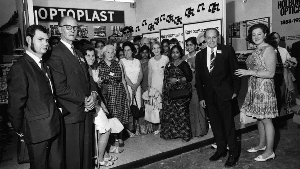 A black and white photograph of a group of professionals gathered in a booth with branding for Optoplast. Fashions reflect the time in 1937.