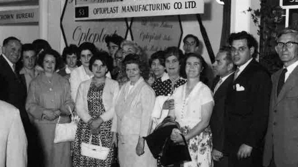 A slightly blurred black and white photo from the 1950s of a group, mostly of women, dressed professionally in front of a booth for Optoplast Manufacturing
