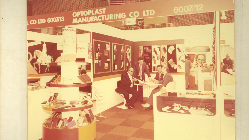 An old photo, captured in faded and sepia tones, shows an exhibition booth branded as Optoplast. On tables around the space are racks of eyewear. On display boards are materials for cases and ophthalmic frames