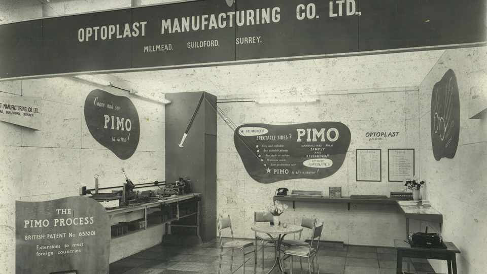 A black and white photograph of an exhibition booth with the Optoplast Manufacturing logo. Signs around the space advertise the patented PIMO machine and process