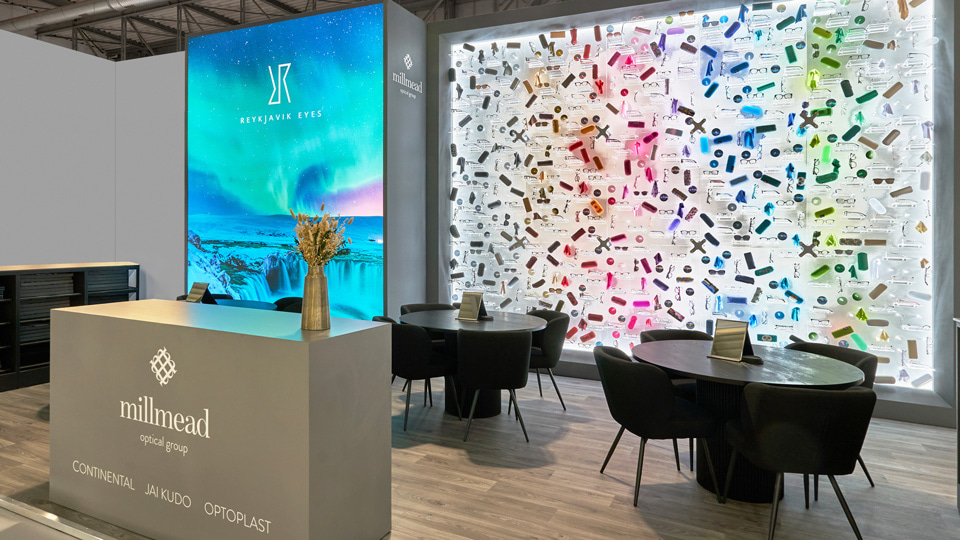 An exhibition stand with large light walls, one displaying an image of the northern lights. A long white light wall is decorated with hundreds of clear shelves, on which are arranged frames, lenses, and cases, in a rainbow of colours from grey to green. Two tables are prepared in the centre