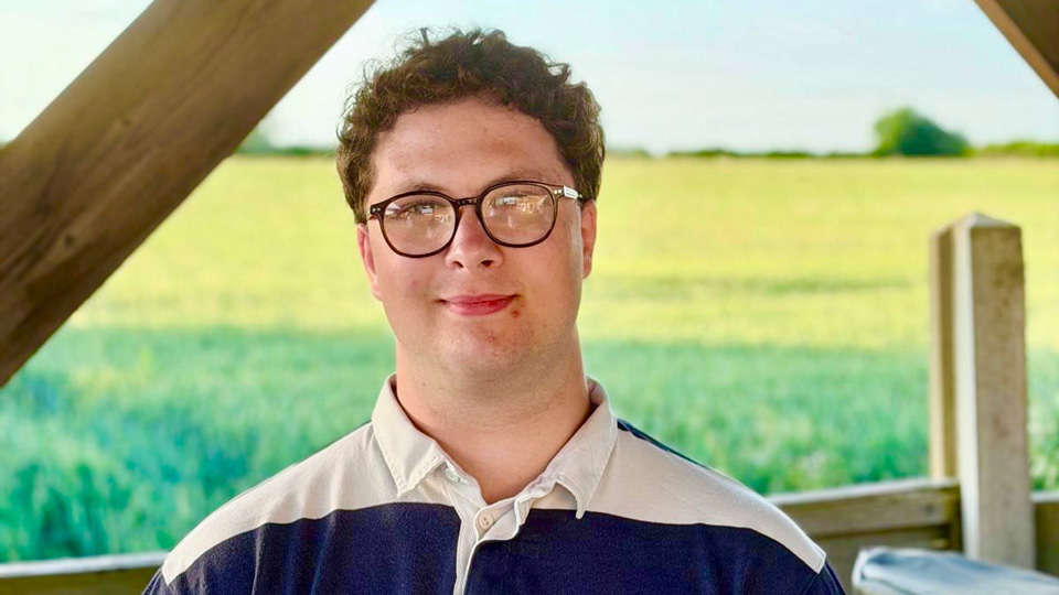 Andrew is photographed in a wooded pergola, which looks out onto a field of green and yellow harvest. He smiles to the camera, wearing rounded glasses, has short brown curling hair, and a rugby style shirt with stripes of white and navy