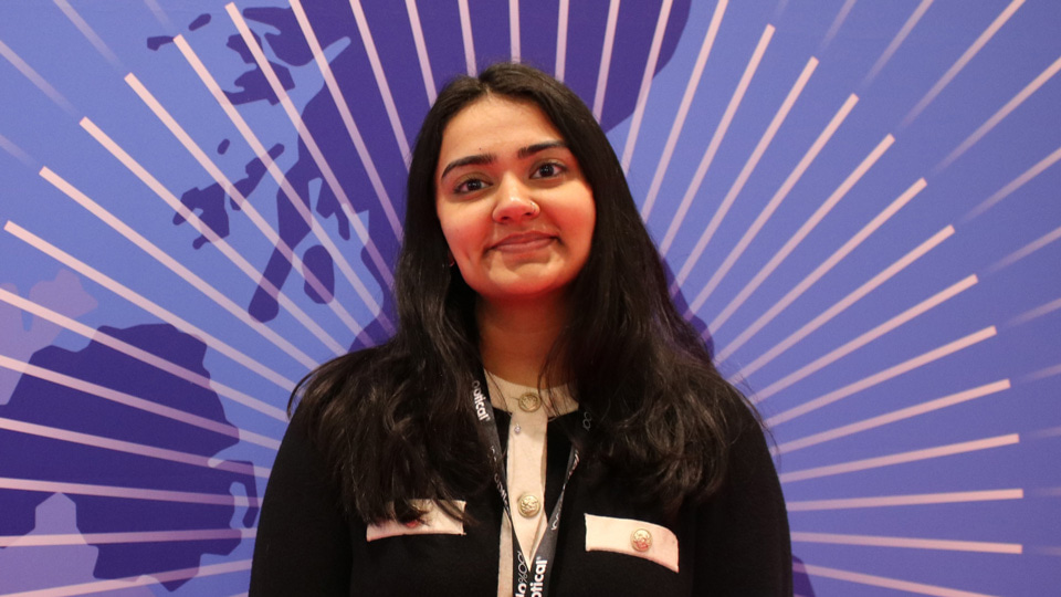 Heer is dressed in professional attire and stands in front of a large poster for the AOP