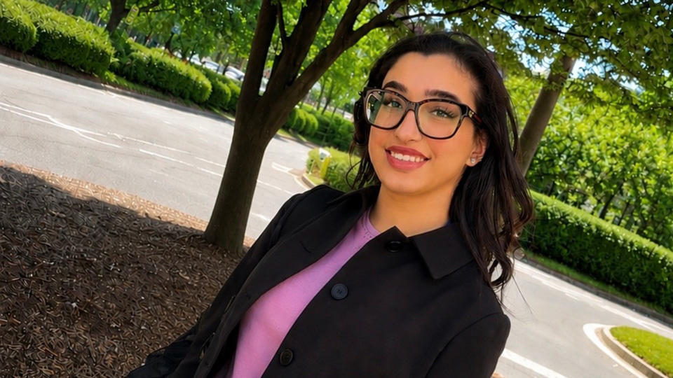 Umamah stands in a bright, green space under tree cover. She has long dark hair past her shoulders and square glasses with a slight lift to the corners. She wears a pink top, black trousers and jacket
