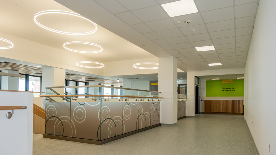 The main staircase and outpatients reception. The stairs are protected by frosted glass with a circle ripple pattern in white and black, reflecting a similar ring lighting above. In the distance is a wood pannelled reception desk with bright green wall behind