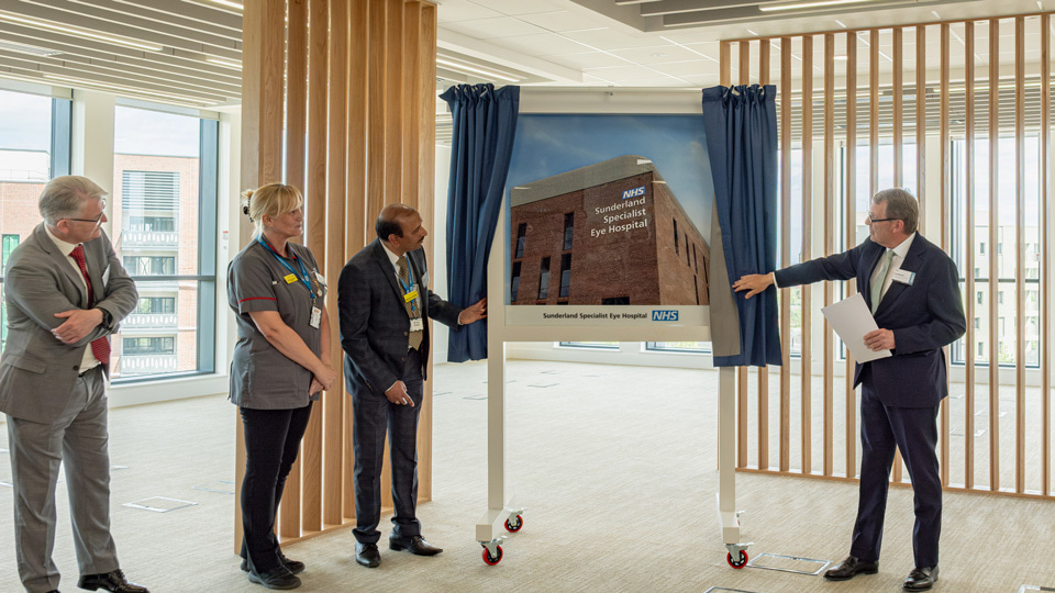 Three men and a woman stand by a display image of the new hospital, blue curtains on either side of the image have been pulled back to reveal the hospital, all in the picture are looking at the image