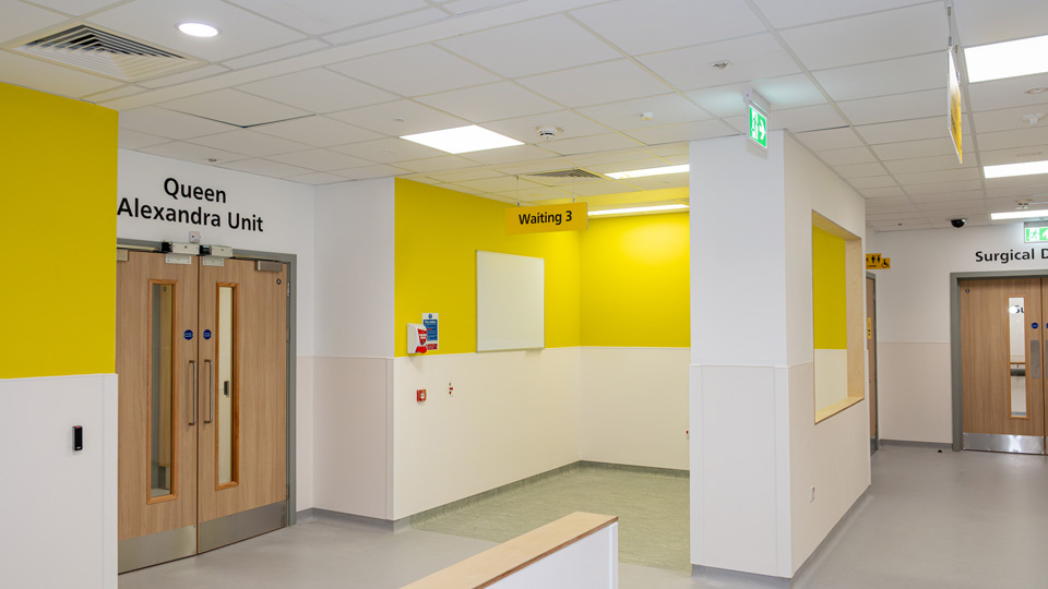 A hospital hallway painting in bright white and yellow. A sign above a double door reads Queen Alexandra Unit