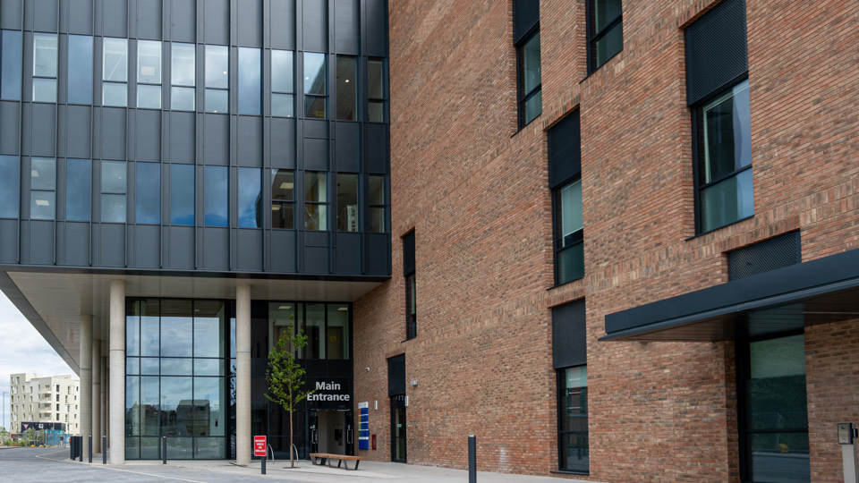 A large brick and glass building with a sign for the Main Entrance, a small tree and bench