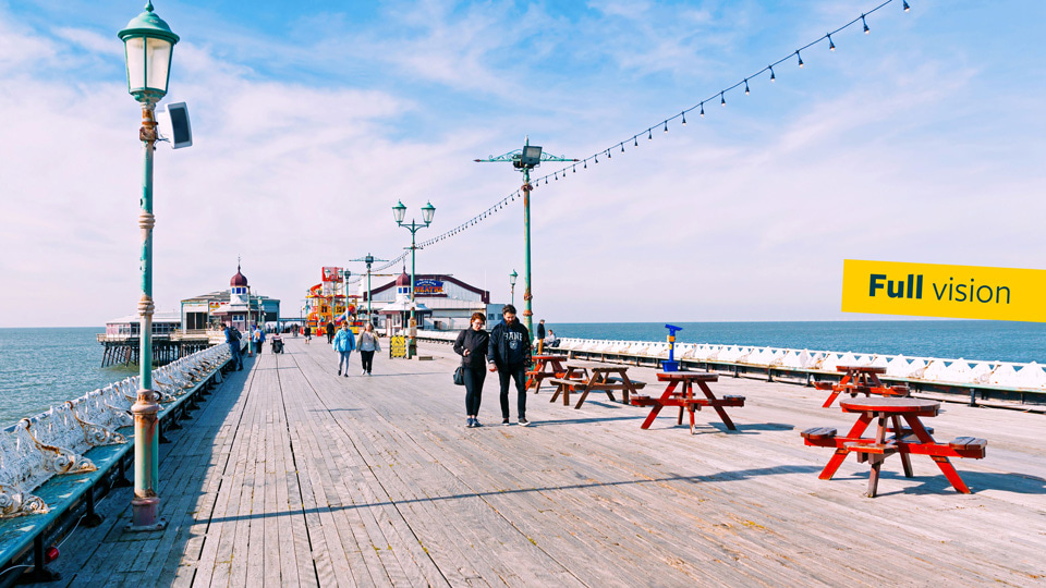 A bright picture of a seaside pier, dotted with red picnic benches, people walking down the centre and buildings at the far end