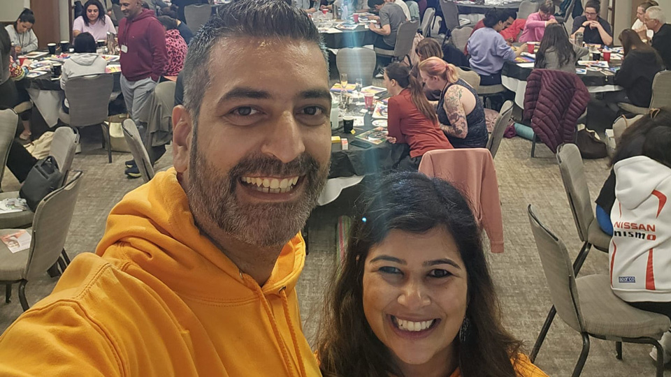 Piyus and Sheena in orange hoodies stand at the front of a room full of large circle tables where participants are painting