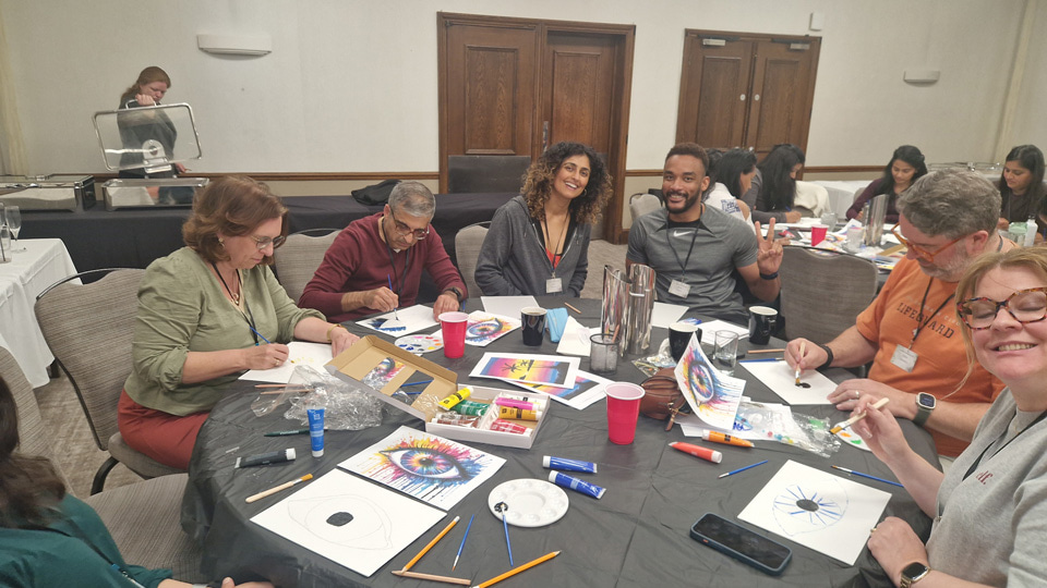 Six people are seated around a large circle table painting from a reference image of a rainbow eye
