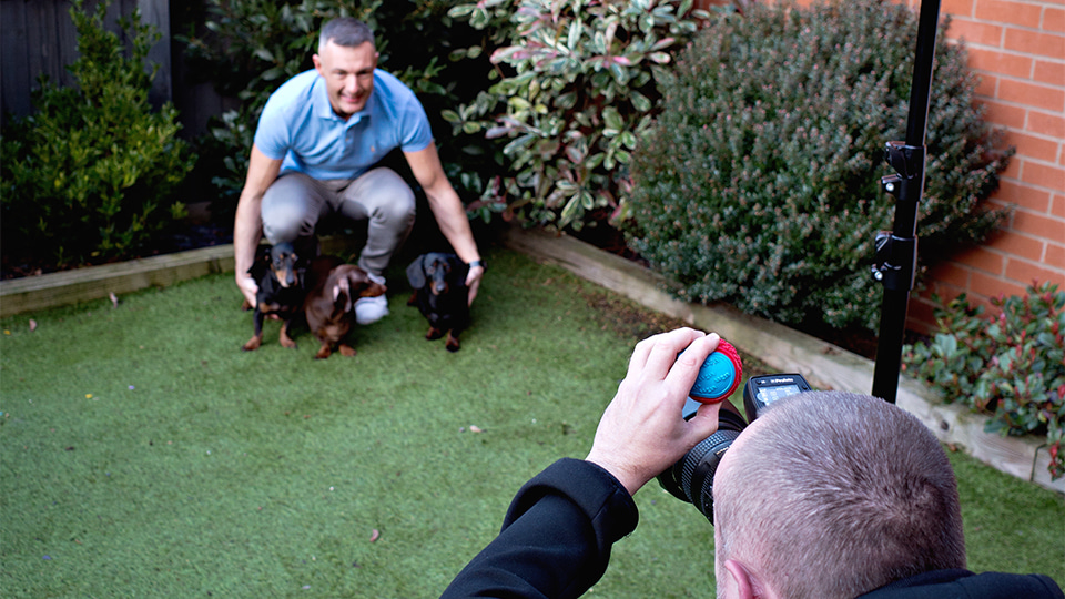 Neil Retallic crouches next to his dogs as he is photographed by a photographer holding a squeaky toy 