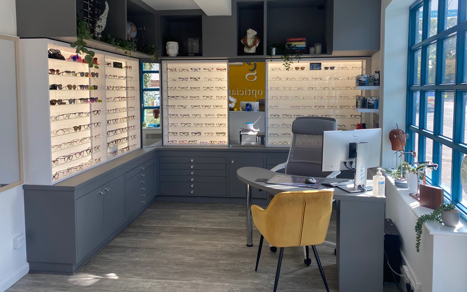 A large window with blue panes has a display of plants and frames on the sill, and shines natural light into the practice. At a dispensing area is a table with a computer, a plush mustard chair and a grey desk chair. The walls have shelves of frames above grey drawers, with grey open cupboards above filled with an array of anatomical, artistic and optical items