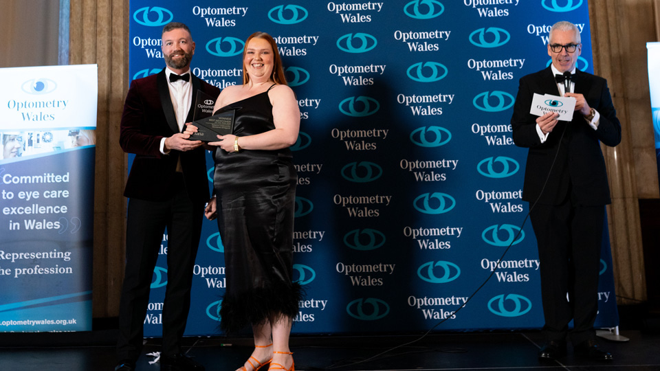 Rebecca wears a long black dress with one strap and a black feathery trim, and bright orange strappy shoes, she smiles as she is handed the award