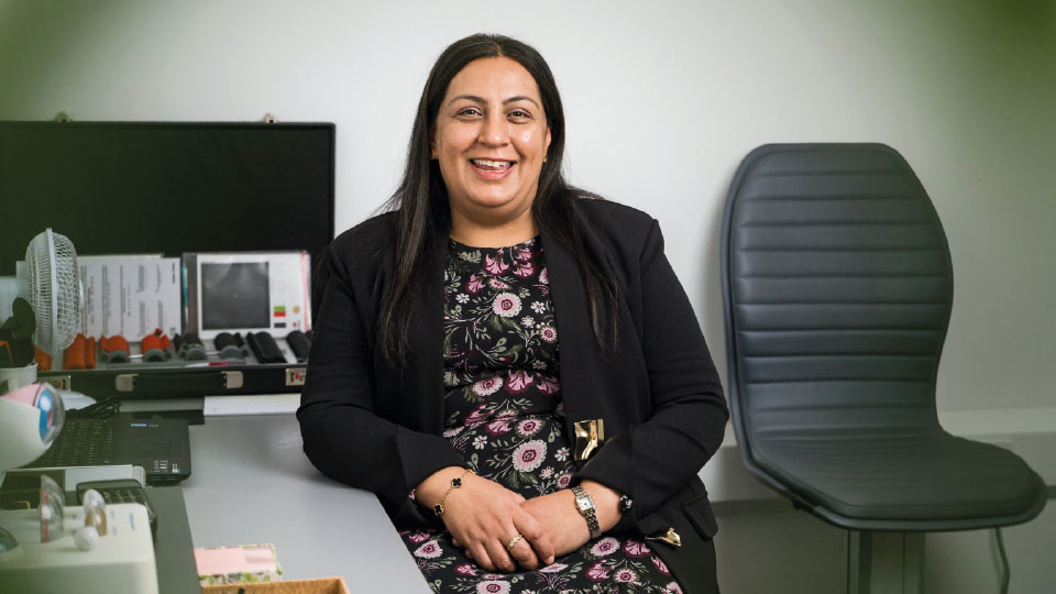 Optometrist Rashida Shaikh smiles at the camera while seated at a desk 