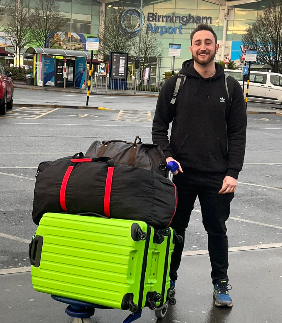 A young man, Luke, standing in front of Birmingham airport with a baggage trolly full loaded with two large suitcases.  