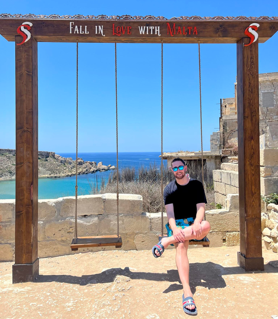 A young man, Luke, is smiling wearing shorts, t-shirt, sandals, sunglasses. He is sitting on a swing on the top of a hill with the view of the ocean behind him. 