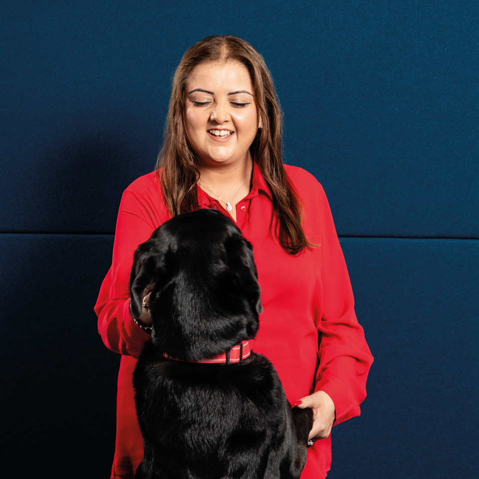 A black labrador wearing a red collar jumps up towards a woman with long dark hair and a red blouse.