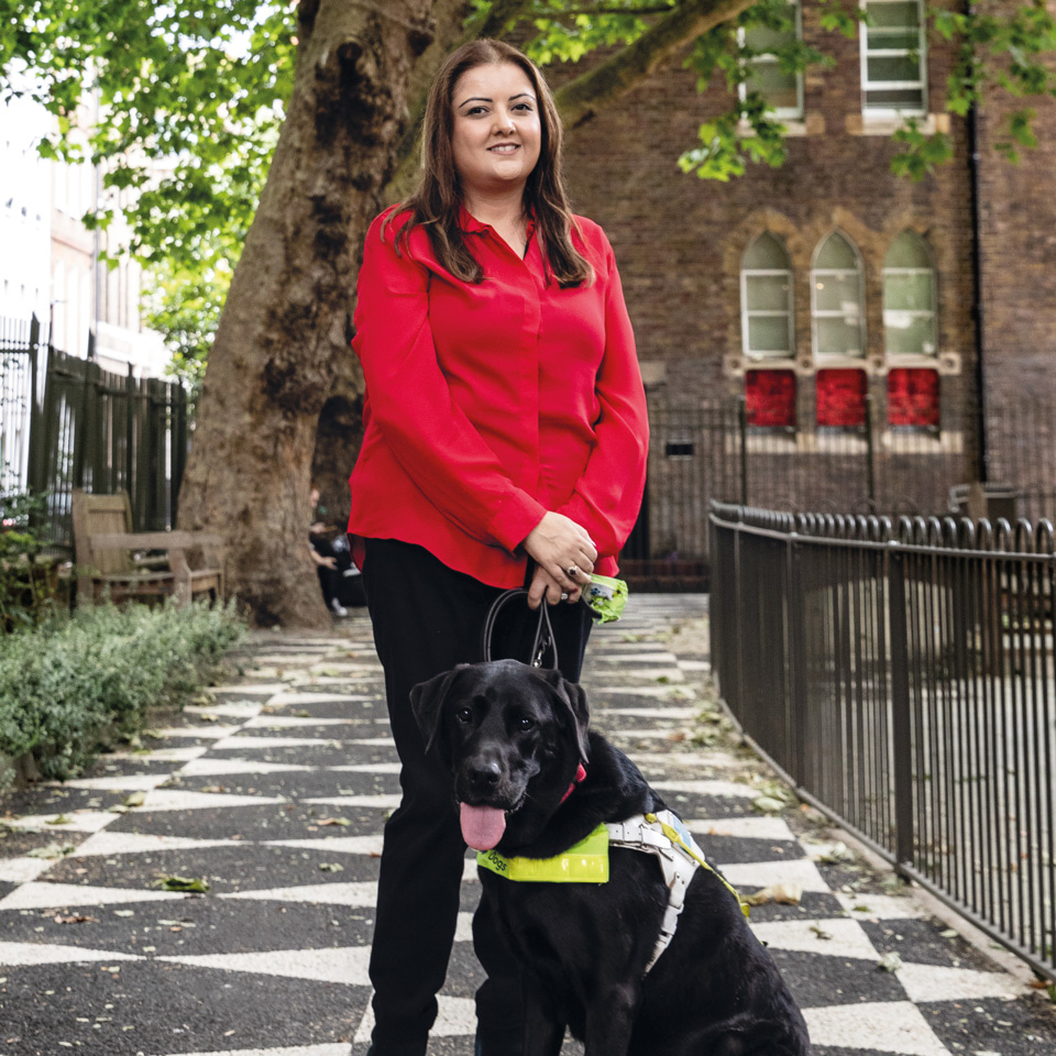 A woman wearing black trousers and a long sleeved red blouse stands outdoors on black and white tiled pavement. Next to her is a black labrador with a fluorescent harness. Behind her there are tall trees and a park railing. 