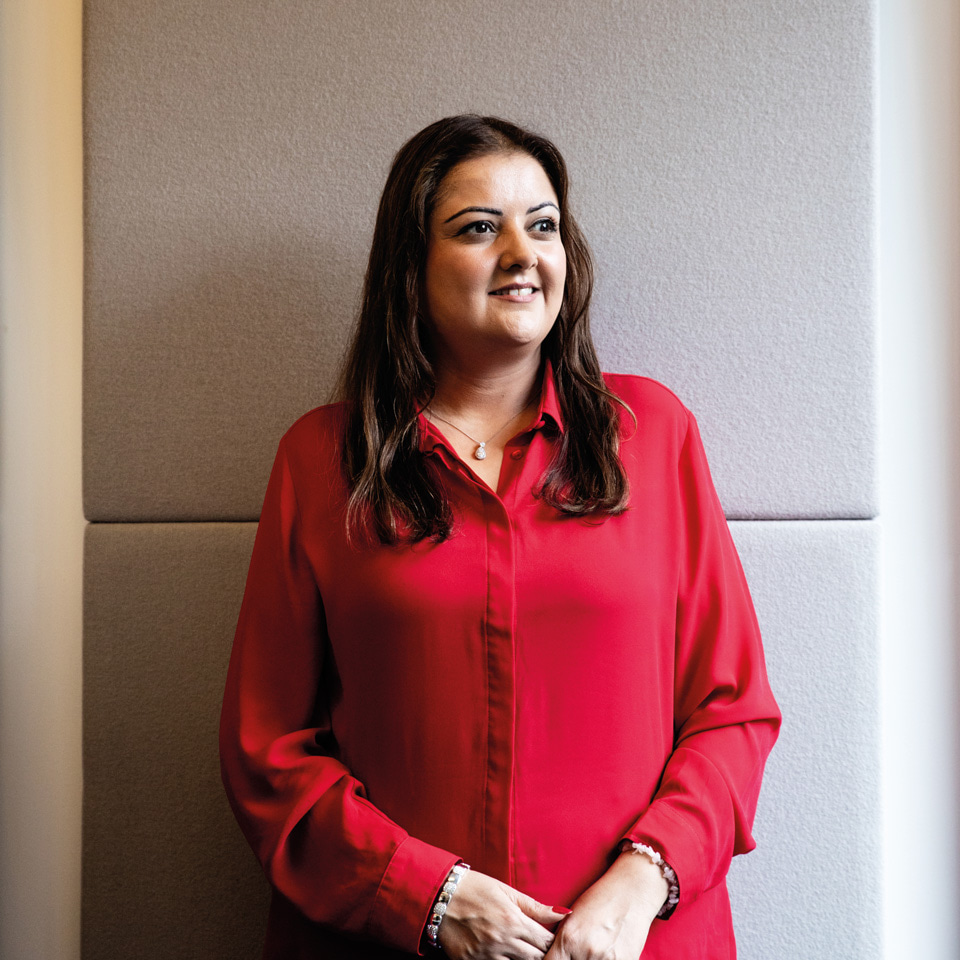 A woman with long dark hair and a longsleeved red blouse stands against a white wall looking to one side. 