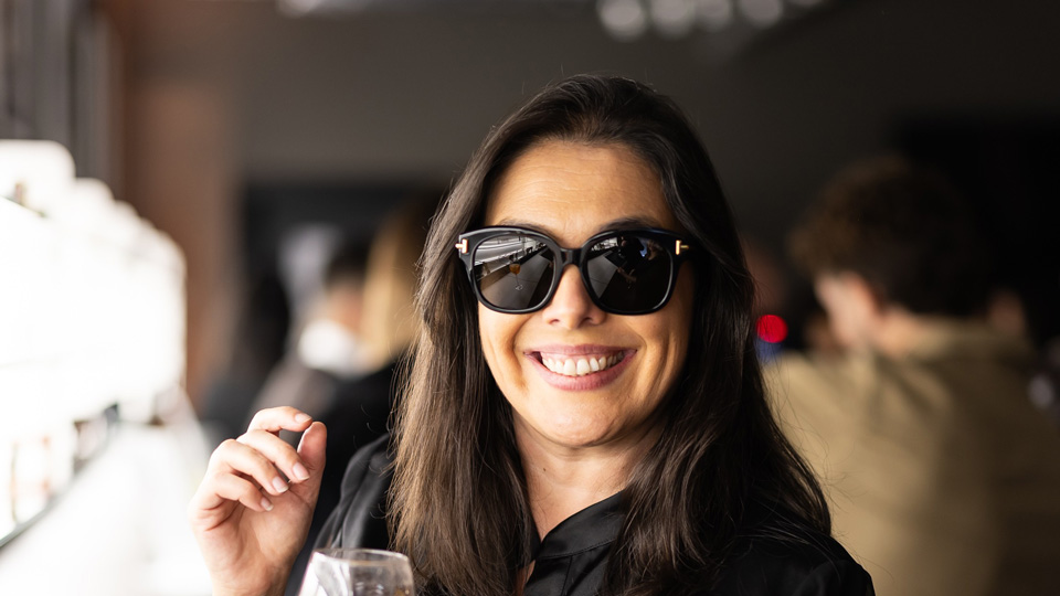 Celsa wears a pair of black sunglasses, standing near a display of eyewear, and a bright smile