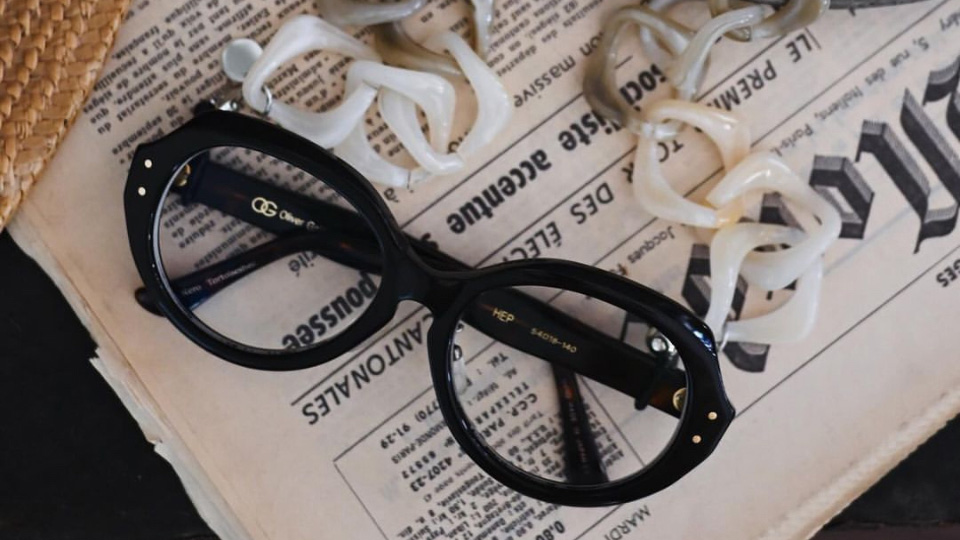 An ombre glasses chain from black to grey to white is attached to a pair of chunky black oval glasses. This is laid out on an old newspaper, a metal plate, and a wicker basket