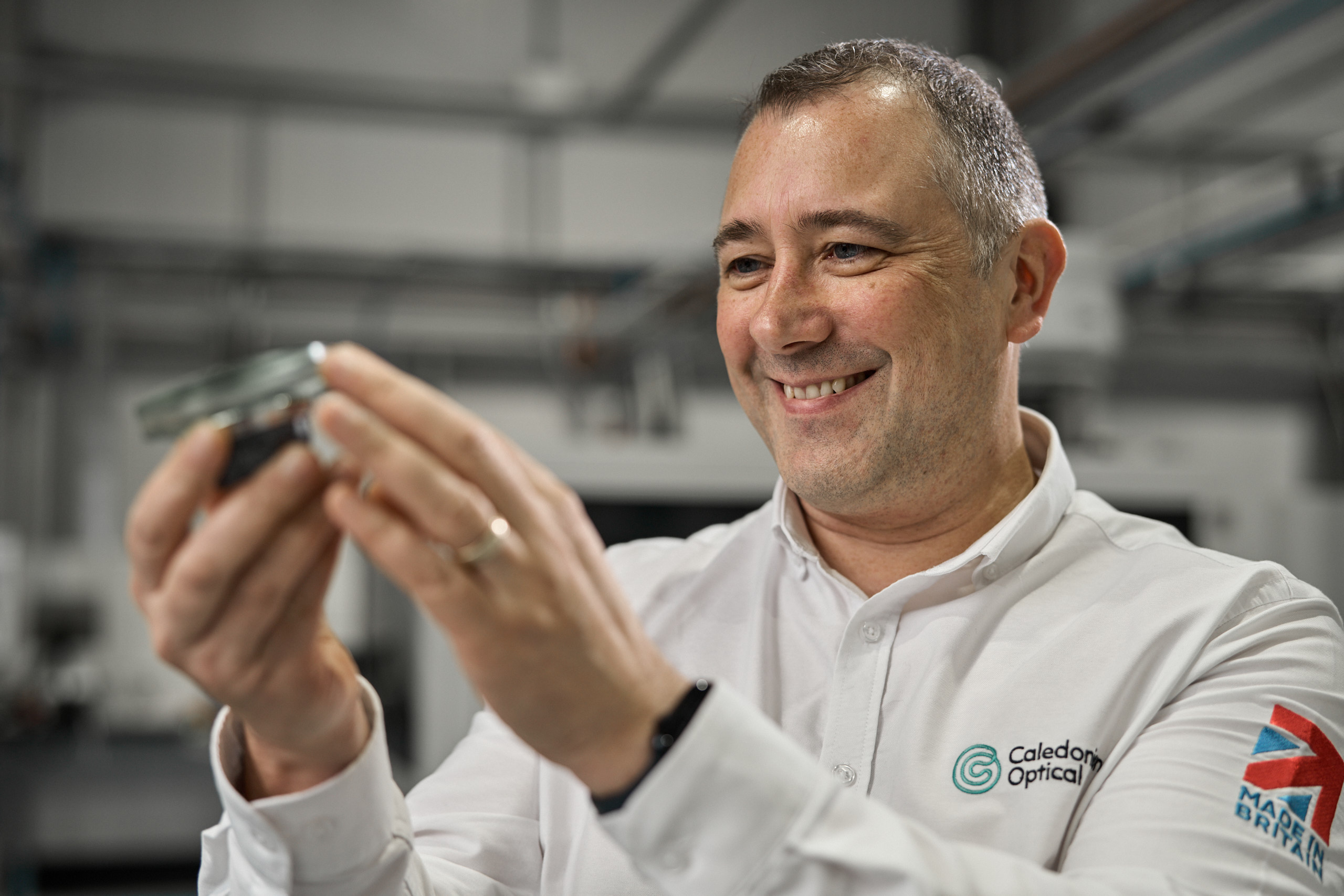 A person in a white shirt with a Caledonian Optical logo holds a lens up, smiling