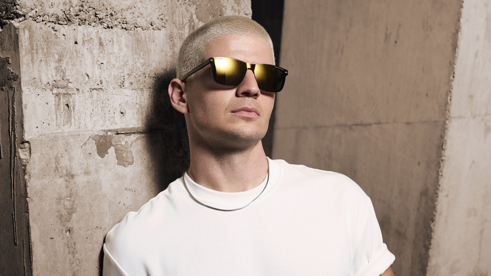 A model with a white tee and blonde buzzcut wears sunglasses with a matte gold lens, catching the light in the top left corners