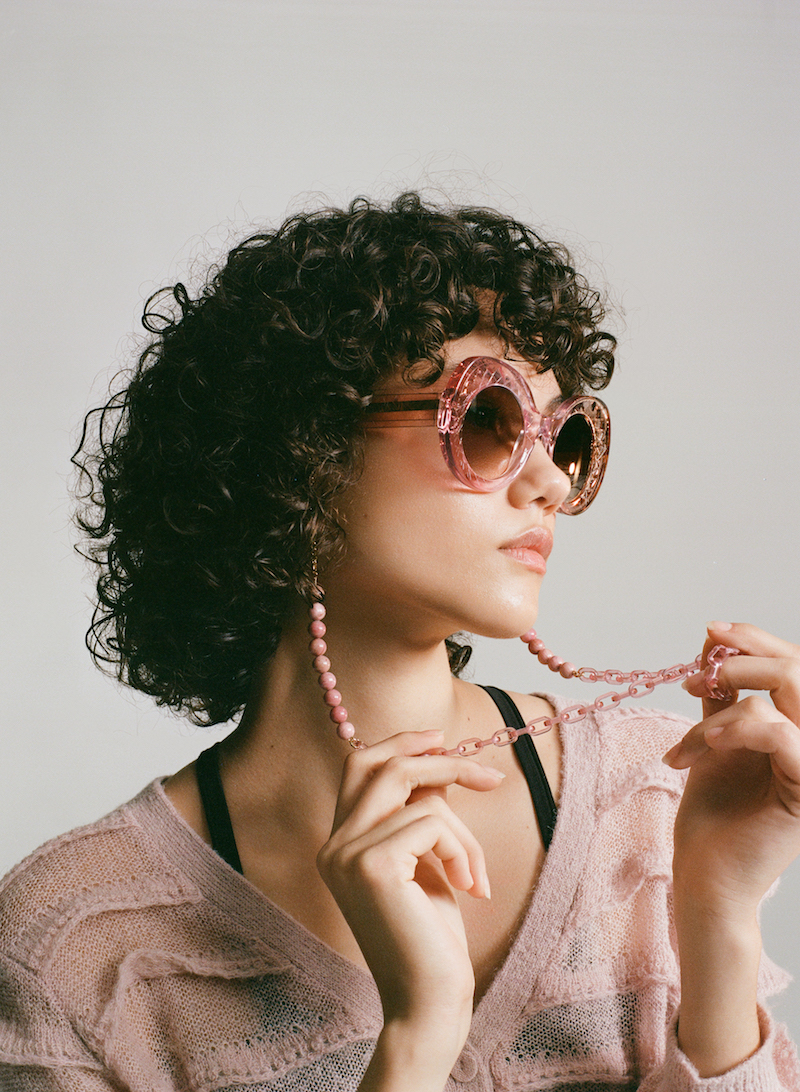 A model looks off camera to the right. She wears oversized butterfly-shaped glasses in a soft transluscent pink with brown lenses and a pink glasses chain that combines beads and chainlink. 