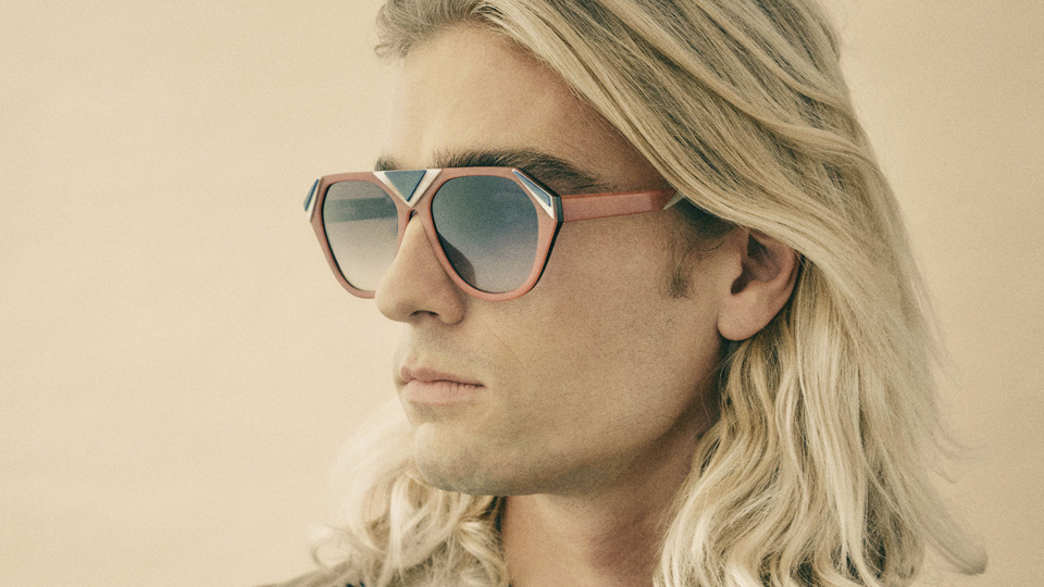A model looks off camera wearing oversized sunglasses in a mask-like shape. The frame is in a red-brown colour, with geometric shapes of white and black. The overall tone of the image is a warm sepia orange