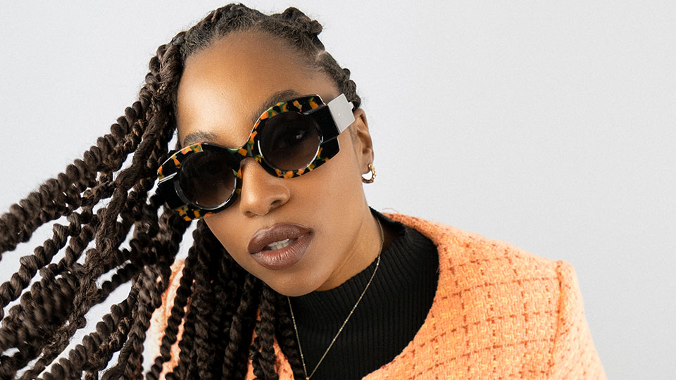 A model wears brown and black sunglasses in a chunky rounded square shape, with thick rectangle arms