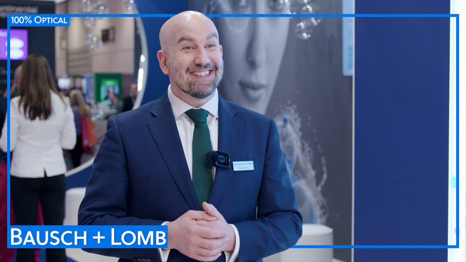 A smiling Richard Smith in a deep blue suit and blue-green tie on the Bausch + Lomb stand at 100% Optical