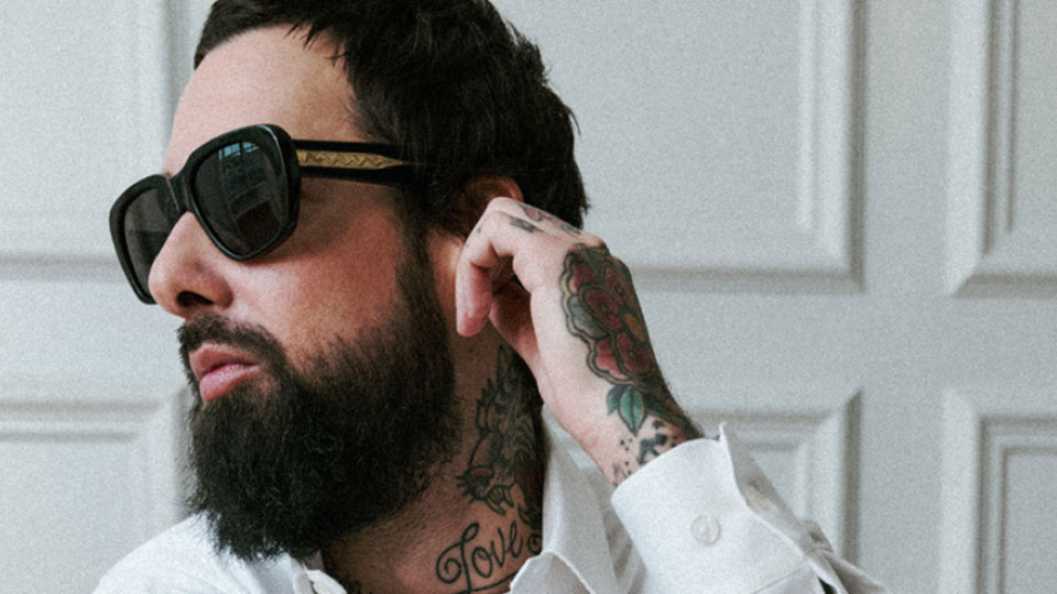 A bearded man wears chunky black sunglasses. The frames have wide arms and a visible core of a patterned gold metal