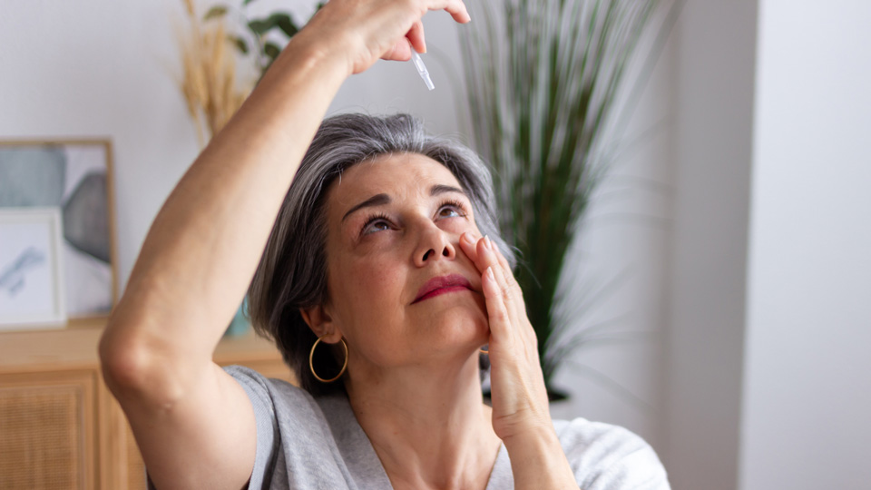 Middle aged woman with grey hair applying eye drops whilst suffering from cataract and glaucoma 