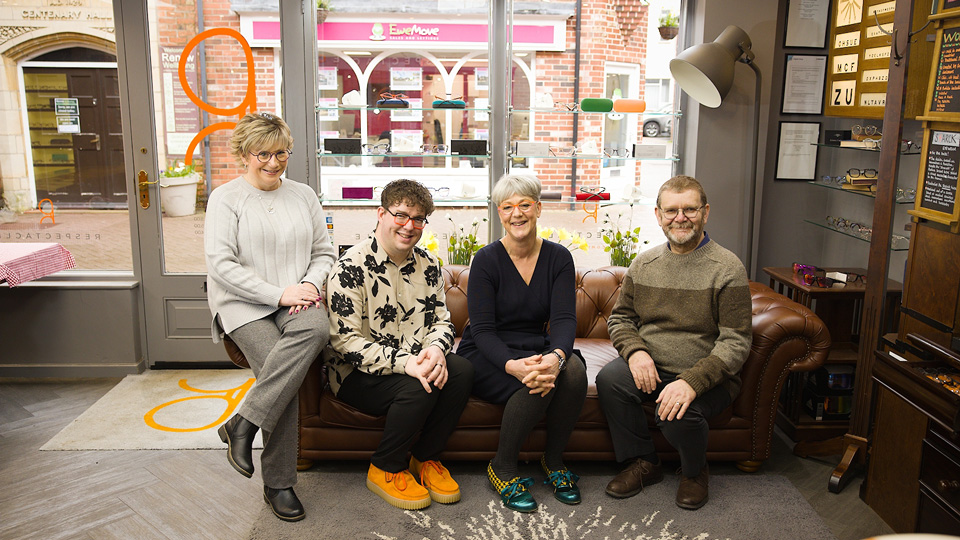 The four-strong practice team sit on a brown leather sofa positioned in the winow of the practice, which looks out onto a quiet High Street. The team have funky eyewear, and several are wearing bright shoes in orange (matching the practice logo on the door) and teal.