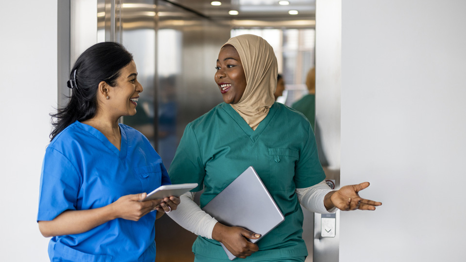 Two clinicians walk down the corridor talking. They are both wearing scrubs and one is wearing a hijab