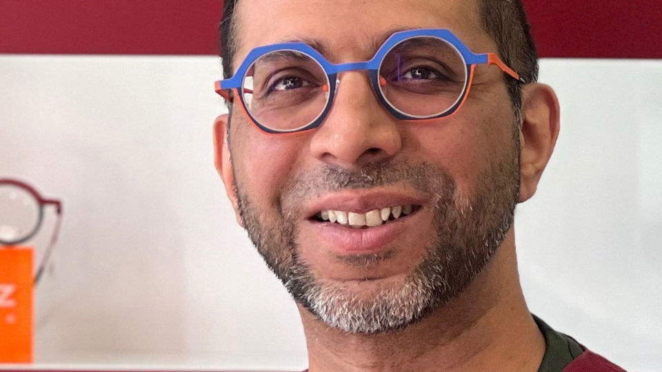 Mohamed Kasmani wears bright geometric spectacles combining blue and orange, and a maroon clinical top