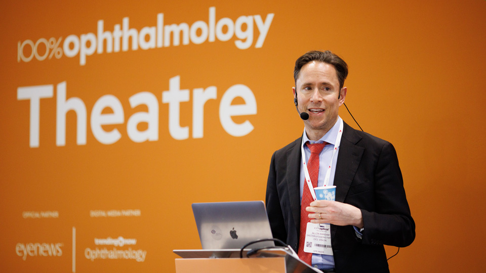 A standing in front of a bright orange background with the '100% Ophthalmology' branding in white font. He is standing at a lectern with a microphone headset, dressed in suit with a red tie.
