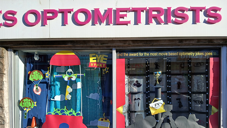 A practice window with paper cut-outs in the style of Toy Story, renamed as ‘Eye Story’. Features include a rocket-shaped claw game which is grabbing alien-green sunglasses