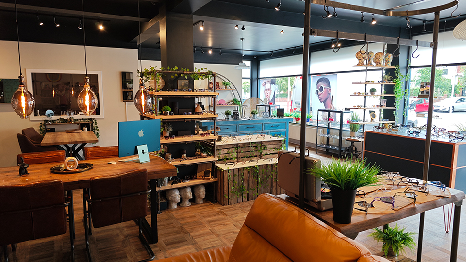 A modern optometry practice with large windows, lots of mid-century style furniture of curving shelves and warm polished woods. A large brown leather sofa is visible. Frames are positioned around the room