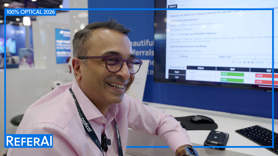 Trusit Dave smiles broadly, he wears a pink shirt and purple metal glasses. He leans against a desk which has a large screen displaying his referral technology