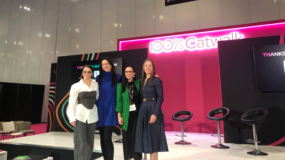 Four women pose on a pink and black 100% Optical catwalk stage. On the left, Hayley wears tinted spectacles, a white shirt and jean corset and dark jeans, beside her is Rebecca who wears a royal blue dress and rimless style spectacles, on her right is Natasha in a black outfit and long green blazer, and finally, Fflur wears a dark navy long sleeved dress