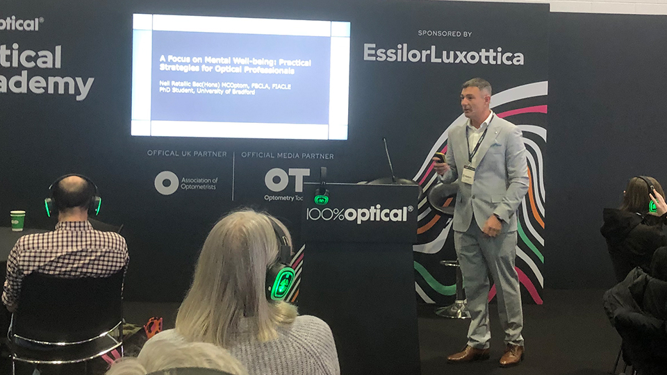 Neil Retallic stands at the front of a seminar space presenting. Audience members are seated around tables wearing headphones