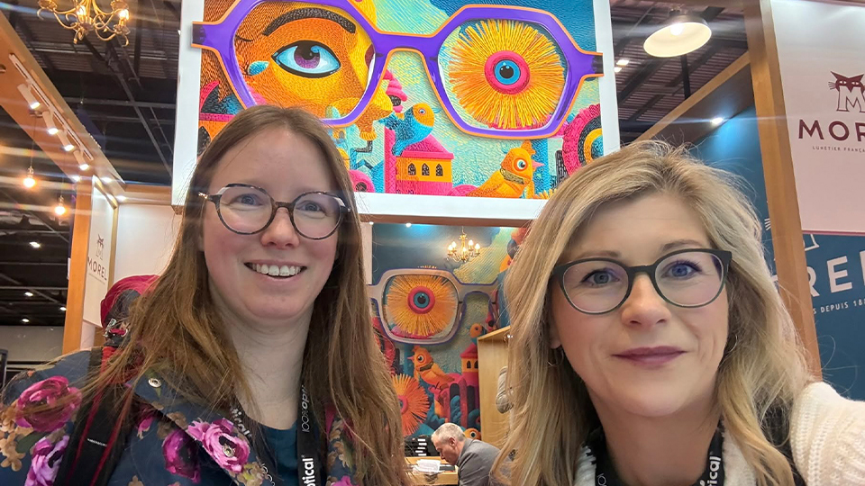 Frances, right, and Laura, left, pose together in front of a vibrant display for Morel Eyewear which features artwork of a pair of bright purple spectacles over an eye and abstract designs