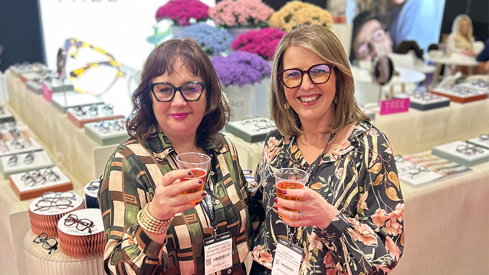 Amanda and her colleague stand together and raise a toast. They stand in front of an exhibition stand for Hudson Eyewear, which has an array of frames on display as well as vases of colourful flowers
