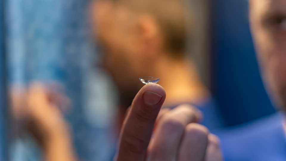 A close up of a contact lens balanced on a man’s finger. He is blurred but appears to be standing in a bathroom